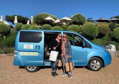 Two women standing next to a blue tour van in the parking lot underneath Mudbrick Vineyard